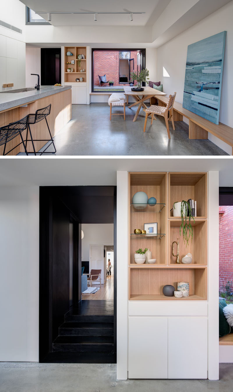In this modern home interior, the dark hallway opens up to the new addition that houses the main social areas. A built-in wood shelf is surrounded by white walls and cabinetry, a strong contrast to the black hallway. #ShelvingIdeas #WoodShelving #OpenPlanLiving