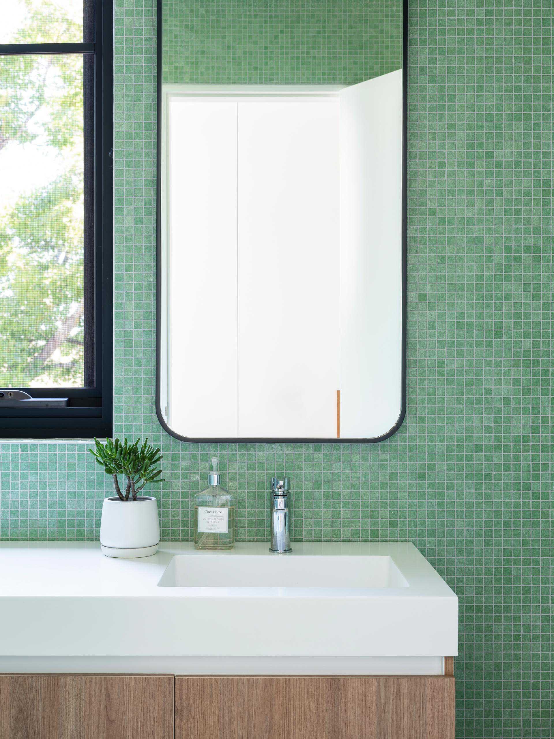 In this modern bathroom, small square green tiles cover the walls, while the black-framed mirror matches the window frame, and the white countertop with integrated sink rests atop a wood vanity.