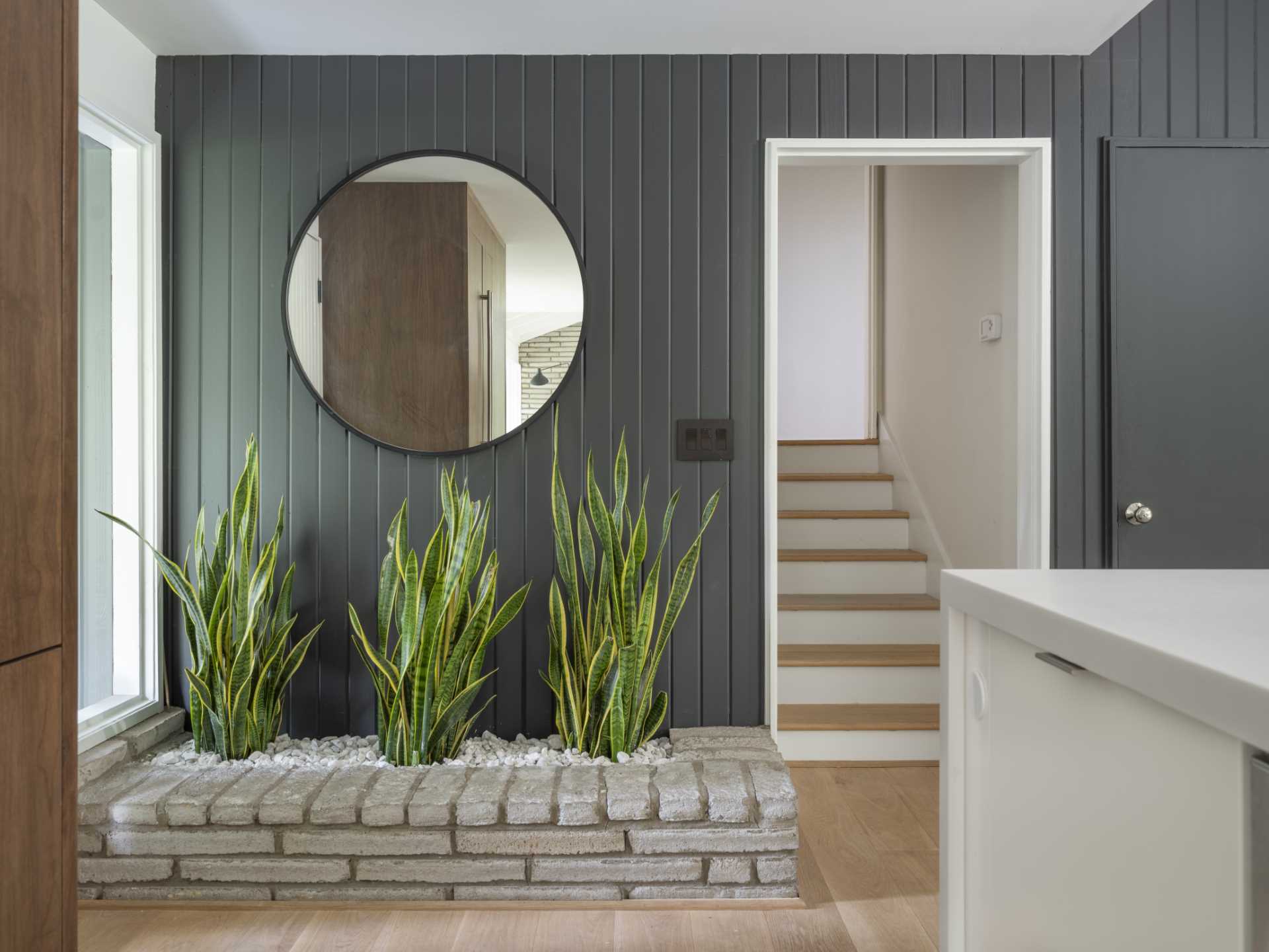 A renovated entryway kept the brick planter by the wall, but now the wood has been painted grey and light wood flooring has been added. A large round mirror helps to reflect light in the space.