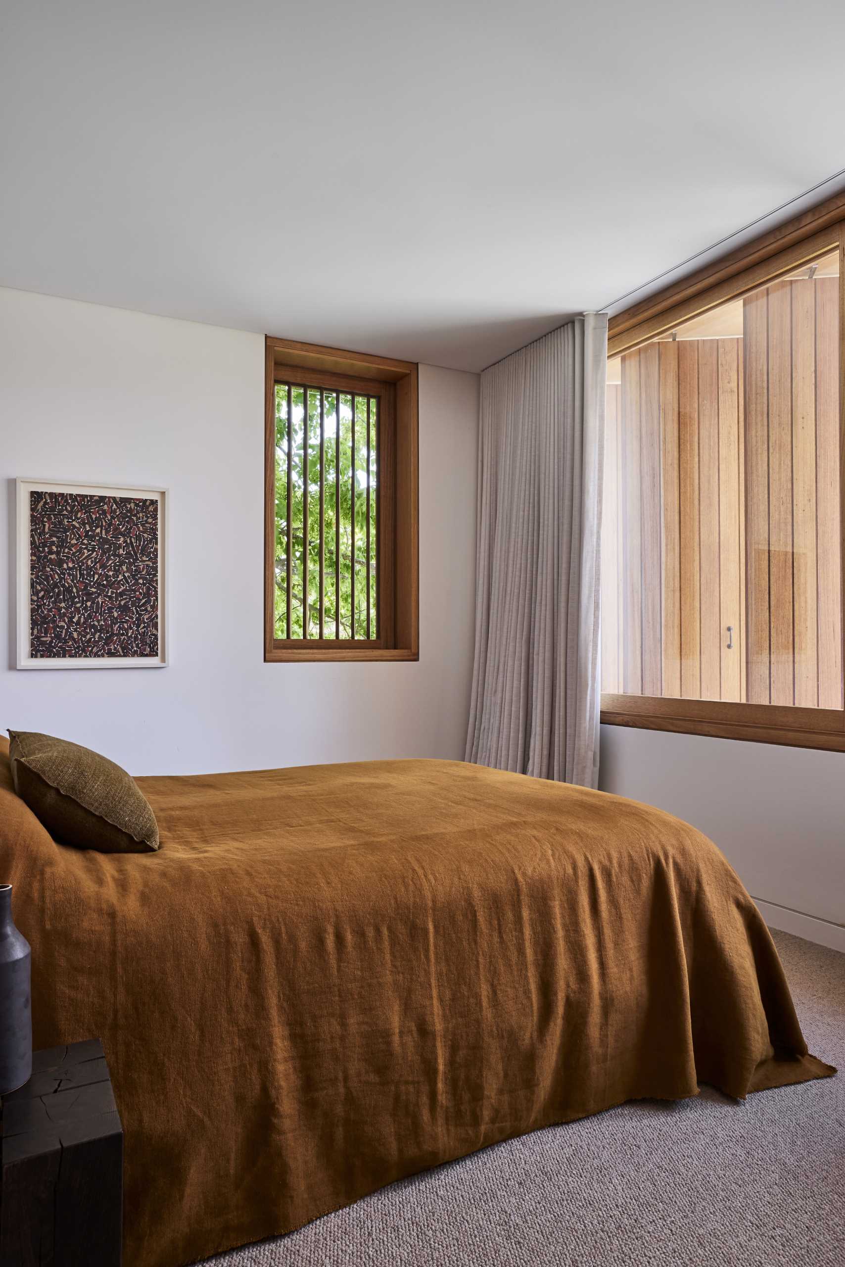 In a bedroom, a large wood-framed picture window is complemented by a smaller window with tree views.