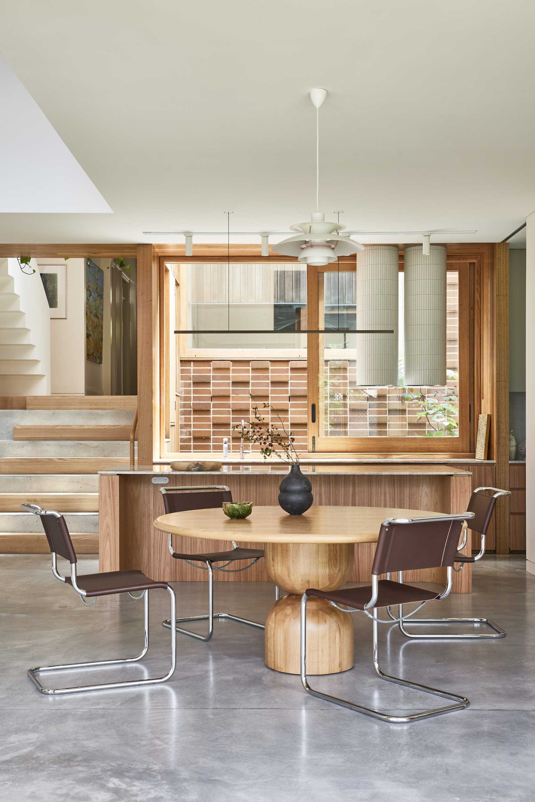 In the new kitchen and dining area, a round dining table is positioned below a pendant light, while the kitchen has an island and a large sliding window that provides views of the original home.