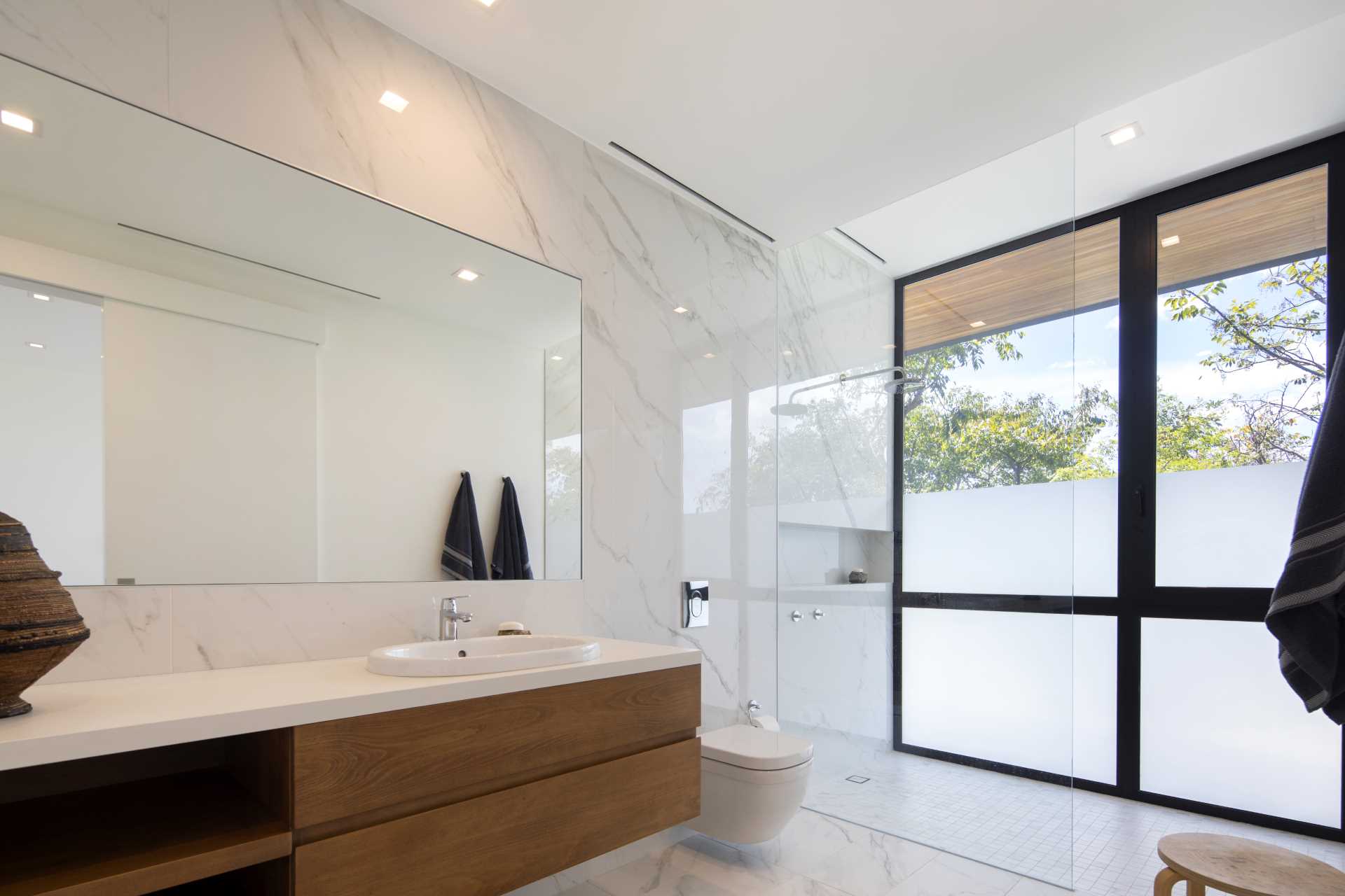 A contemporary bathroom design with a material color palette of wood, light colored tiles, white countertops, and gl،.