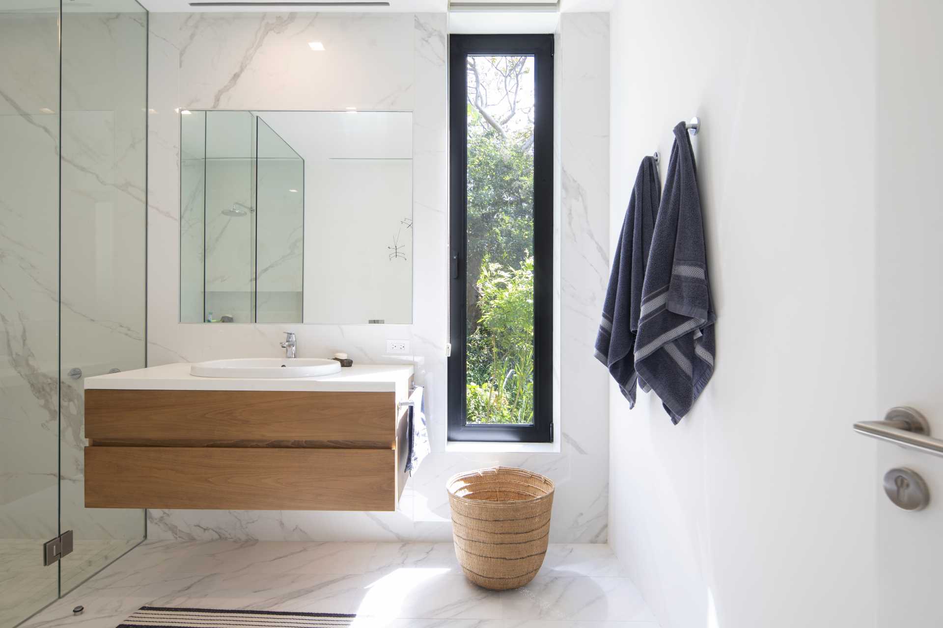 A contemporary bathroom design with a material color palette of wood, light colored tiles, white countertops, and gl،.