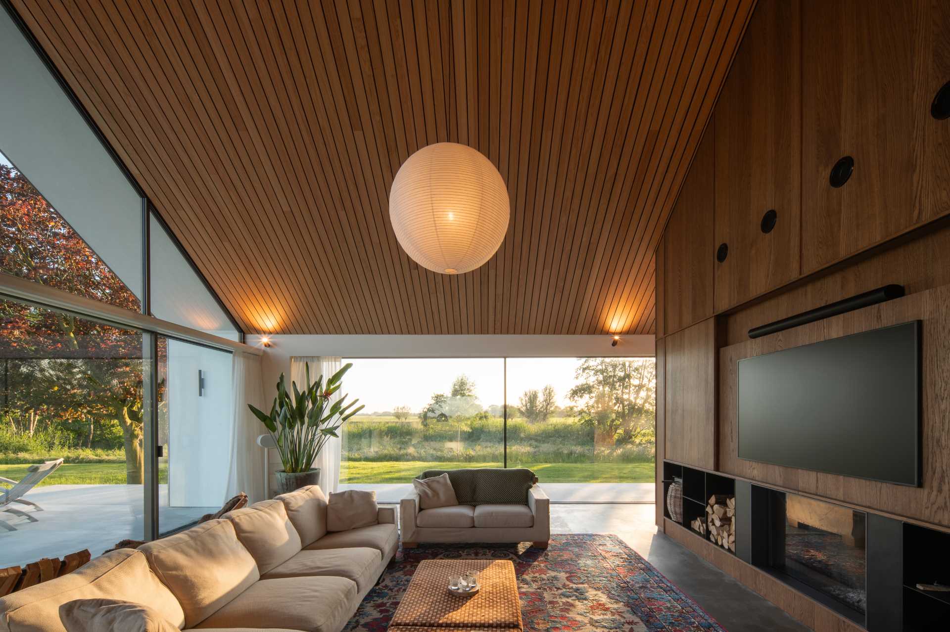 In the living room, it's easy to see the wood-lined gabled ceiling of this modern home. The living room includes sliding doors at one end, a fireplace with wood storage, and a custom shelving unit with a hidden TV.