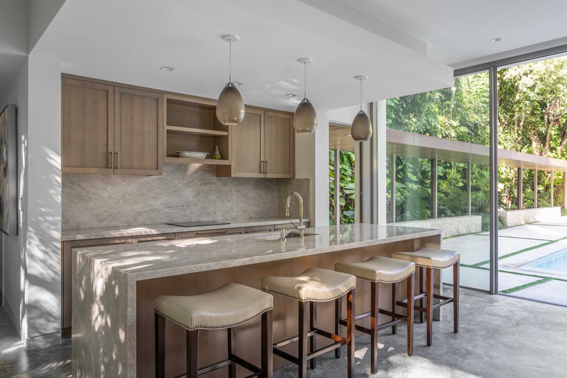 A modern backyard pavilion includes a kitchen with wood cabinetry, an island, and stool seating.