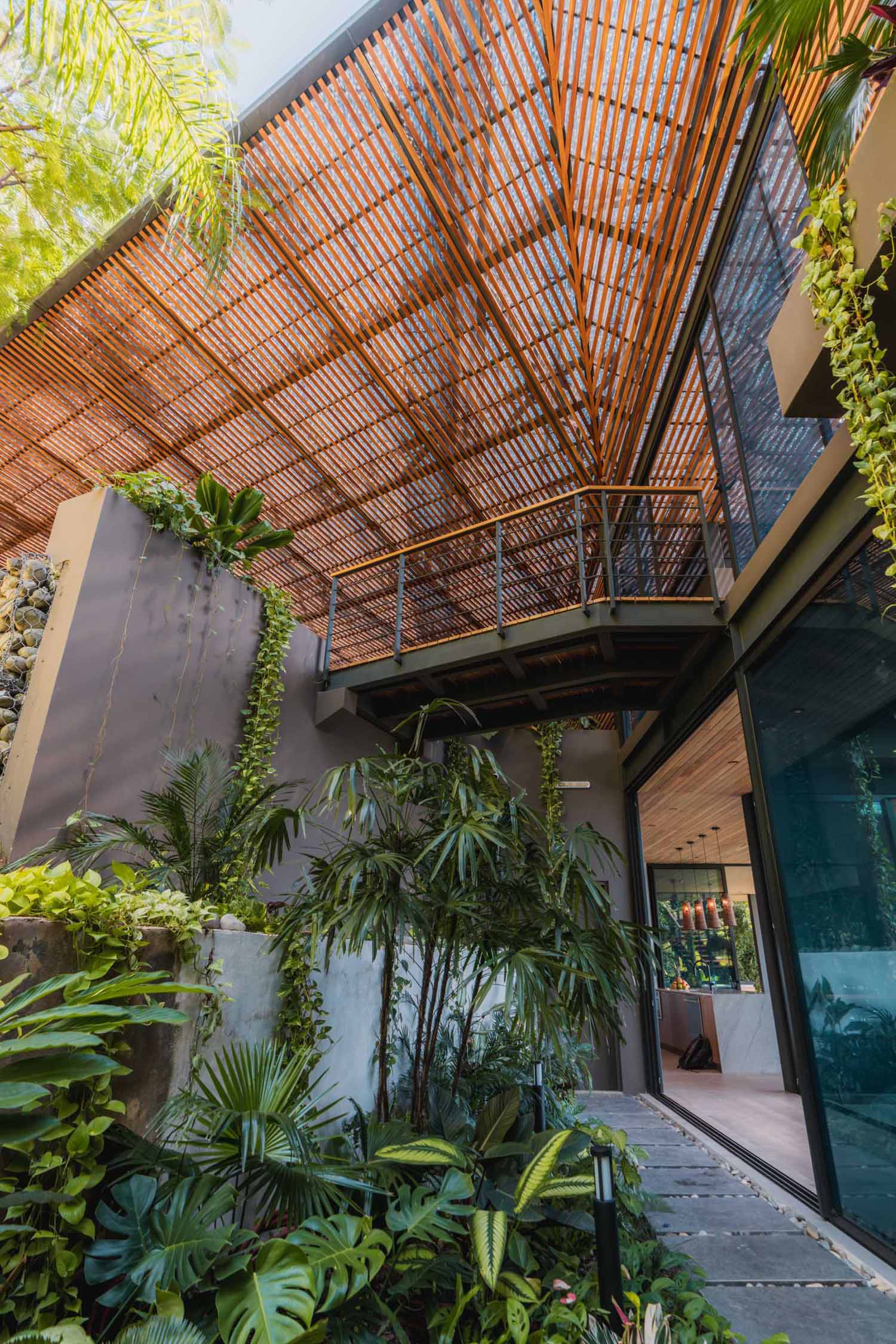 A closer look at the roof of this modern tropical home reveals soft light that permeates through the perforated panels, translucent sheets, and an array of teak louvers, creating an ambiance reminiscent of a dense jungle.