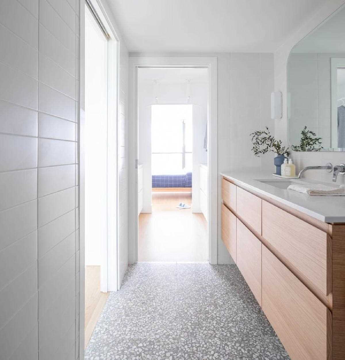 In the bathroom, the Terrazzo tiles make another appearance on the floor, complementing the tiles in the kitchen. A wood vanity adds a natural element and ties in with the bed frame seen through the doorway.