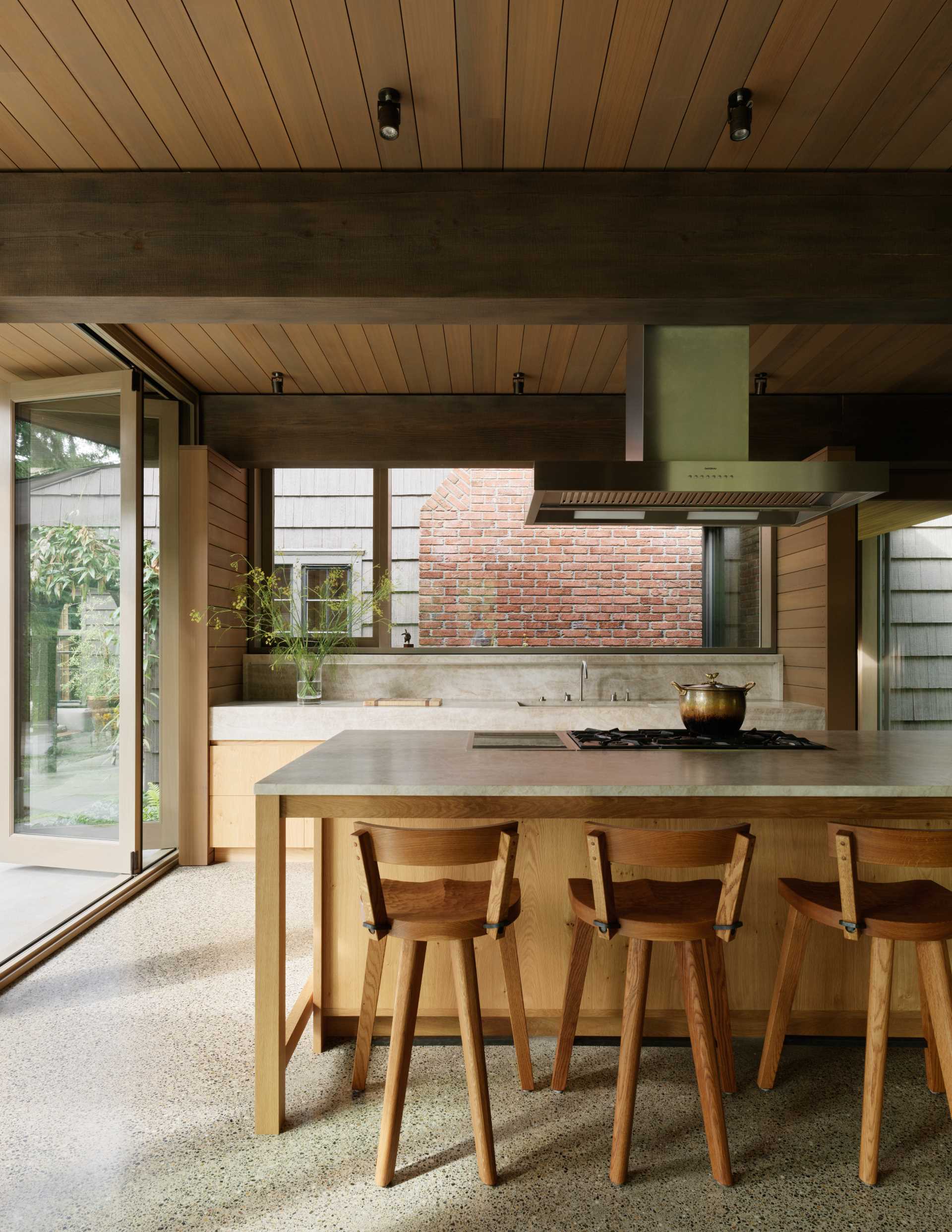 This kitchen interior has a polished concrete floor, stained knotty-oak cabinetry, and Quartzite countertops, including a custom stone kitchen sink. Appliances are fully integrated into the cabinetry. The space is organized around a large island.