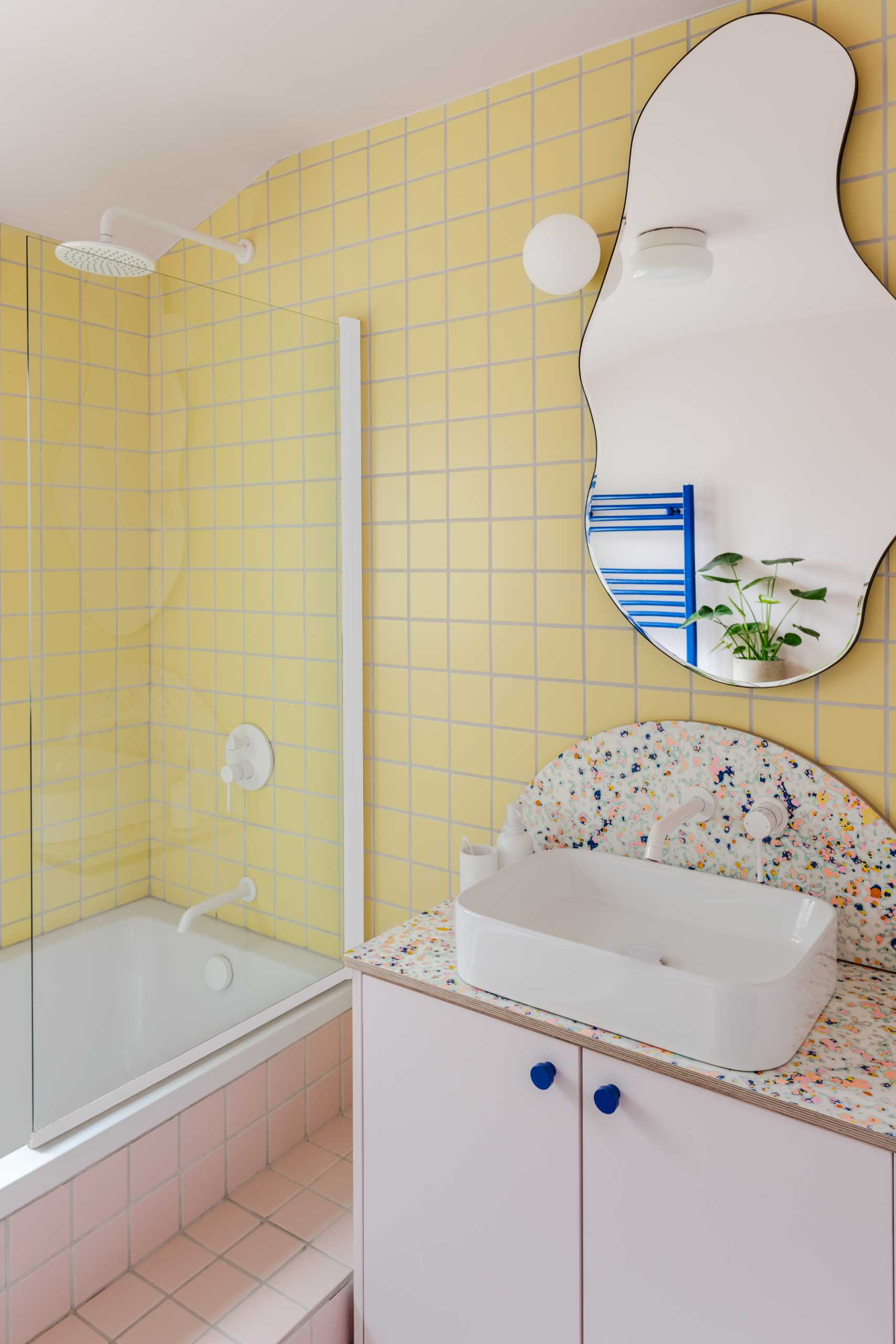 In this bathroom, square yellow tiles cover the walls and meet the pink tiles that wrap around the bathtub, while an organically shaped mirror adds a fun element.