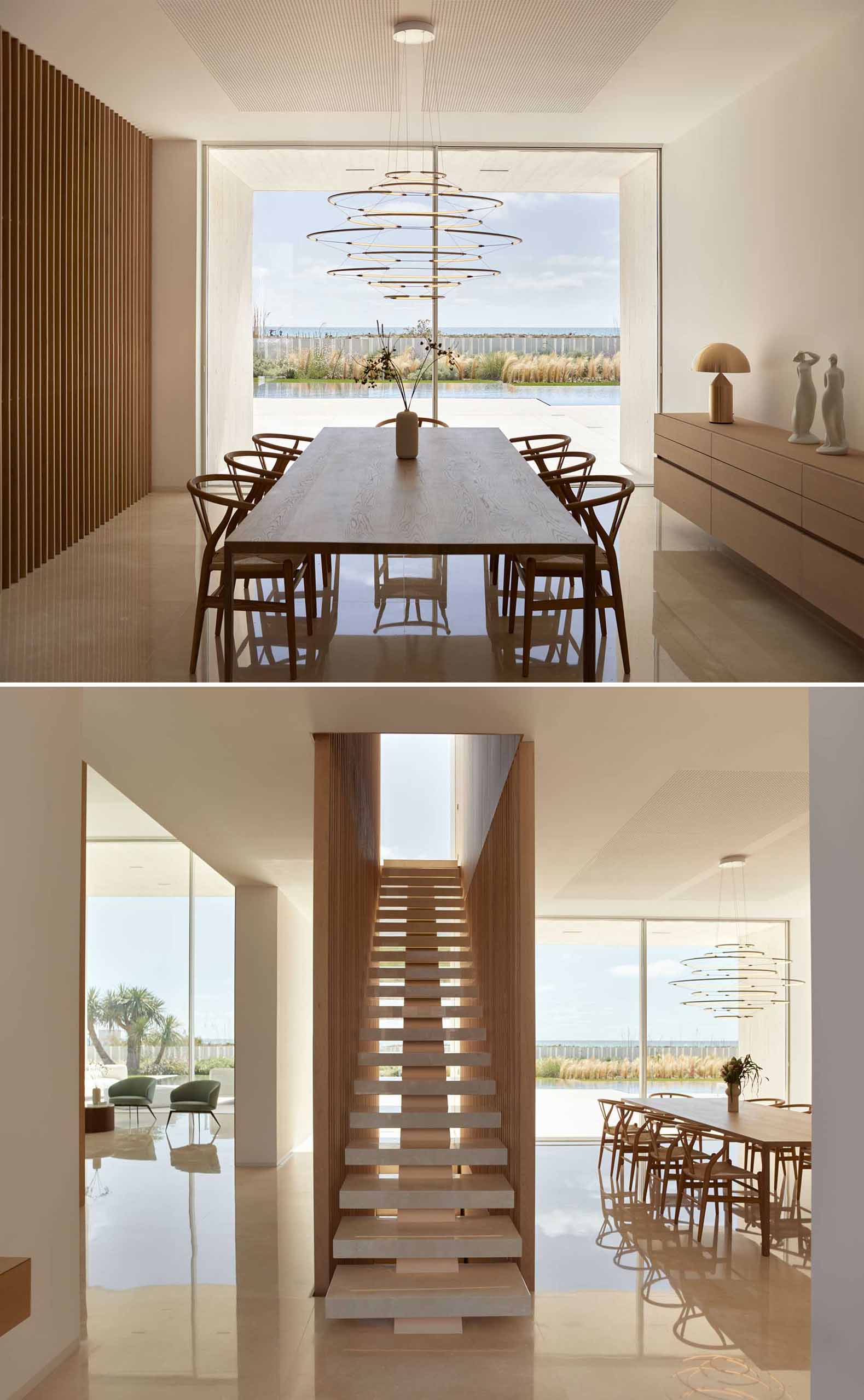 In this dining room, a sculptural light hangs above the table, while windows provide a view of the terrace, pool, and landscape beyond. A wood staircase between the dining room and the living room leads to the bedrooms and bathrooms upstairs.