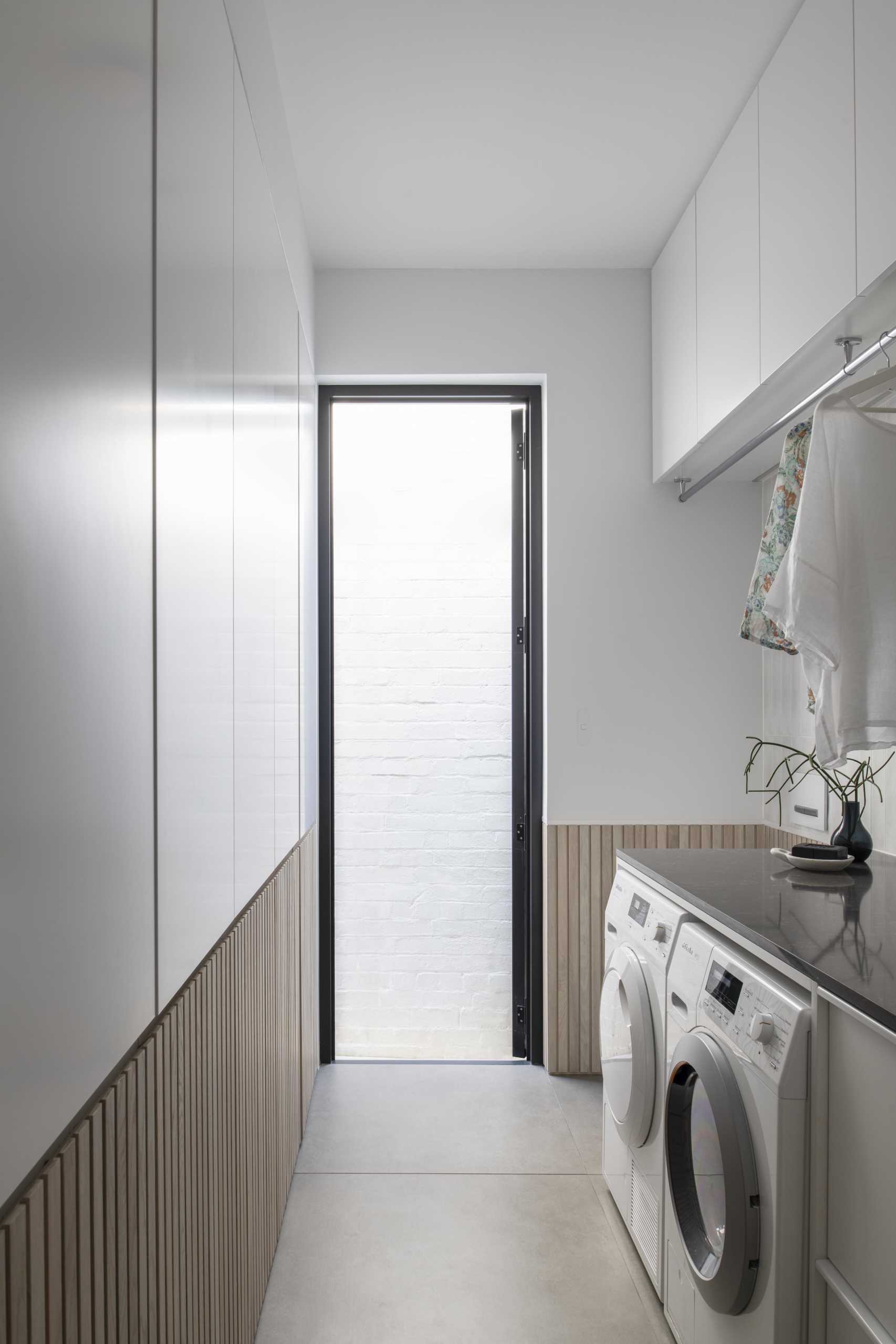 A modern white laundry room with wood paneling that hides three laundry baskets below cabinetry.