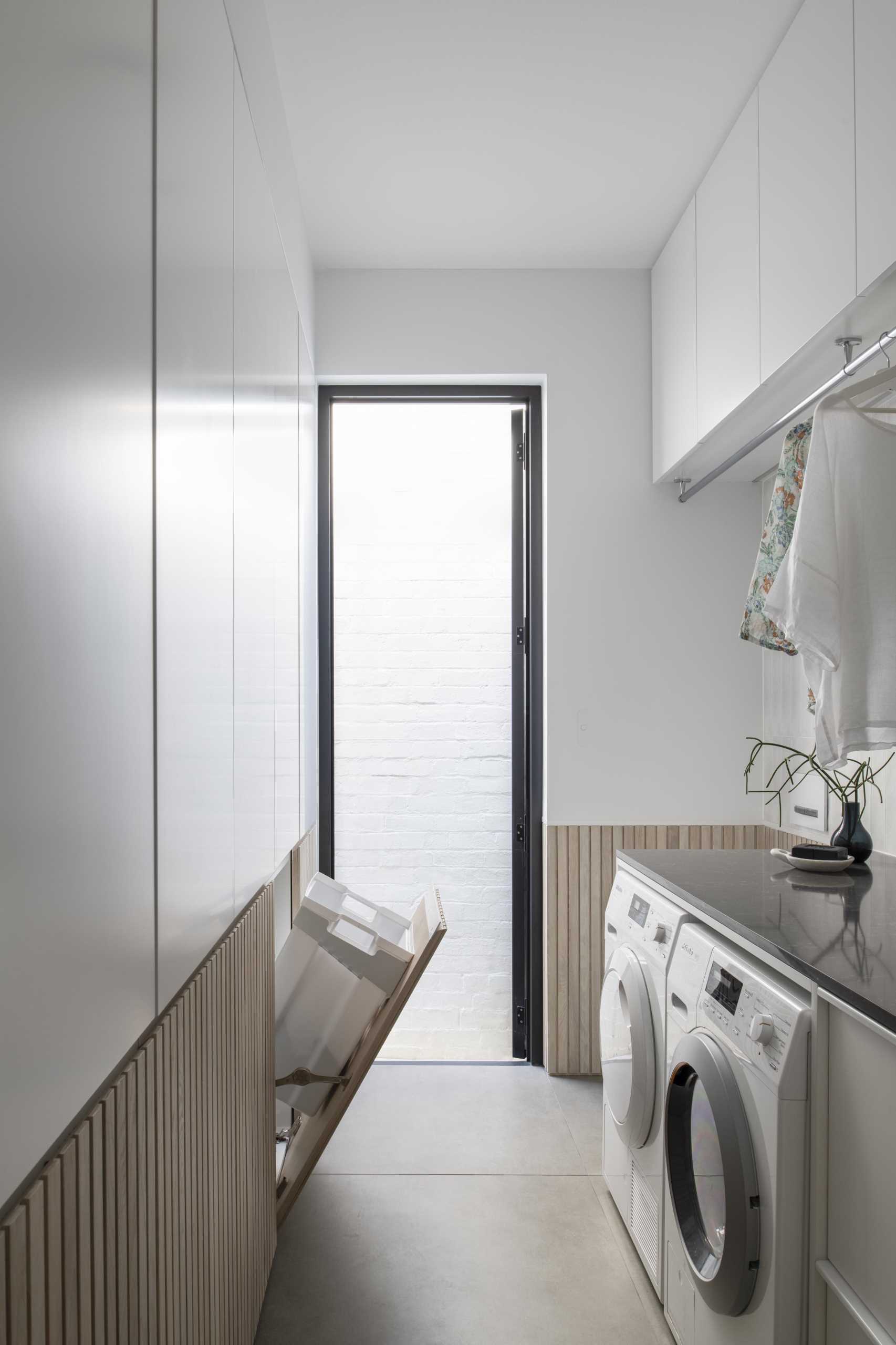 A modern white laundry room with wood paneling that hides three laundry baskets below cabinetry.