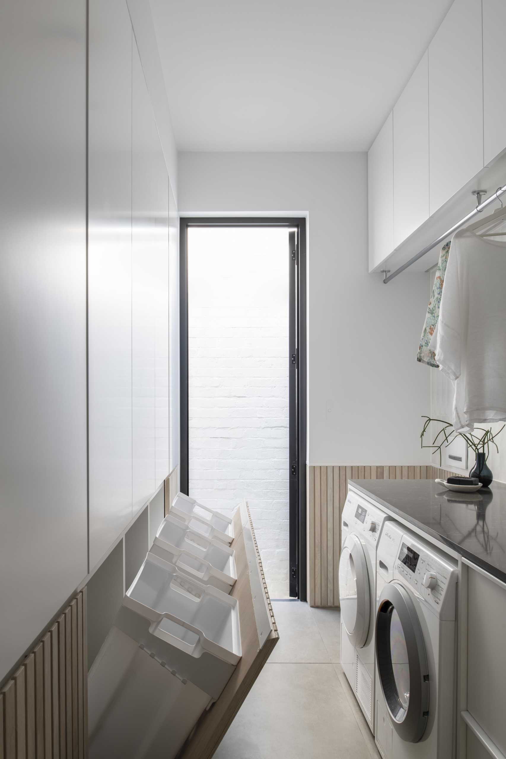 A modern white laundry room with wood paneling that hides three laundry baskets below cabinetry.