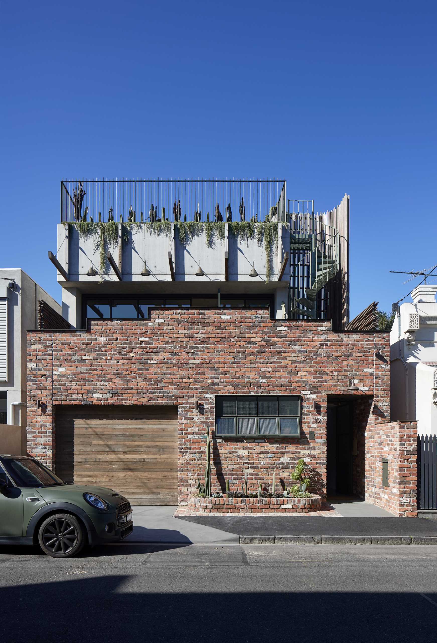 A red brick single-storey facade rises in steps towards the centre, with its pediment topped with a ribbon of steel, concealing the terrace behind. 