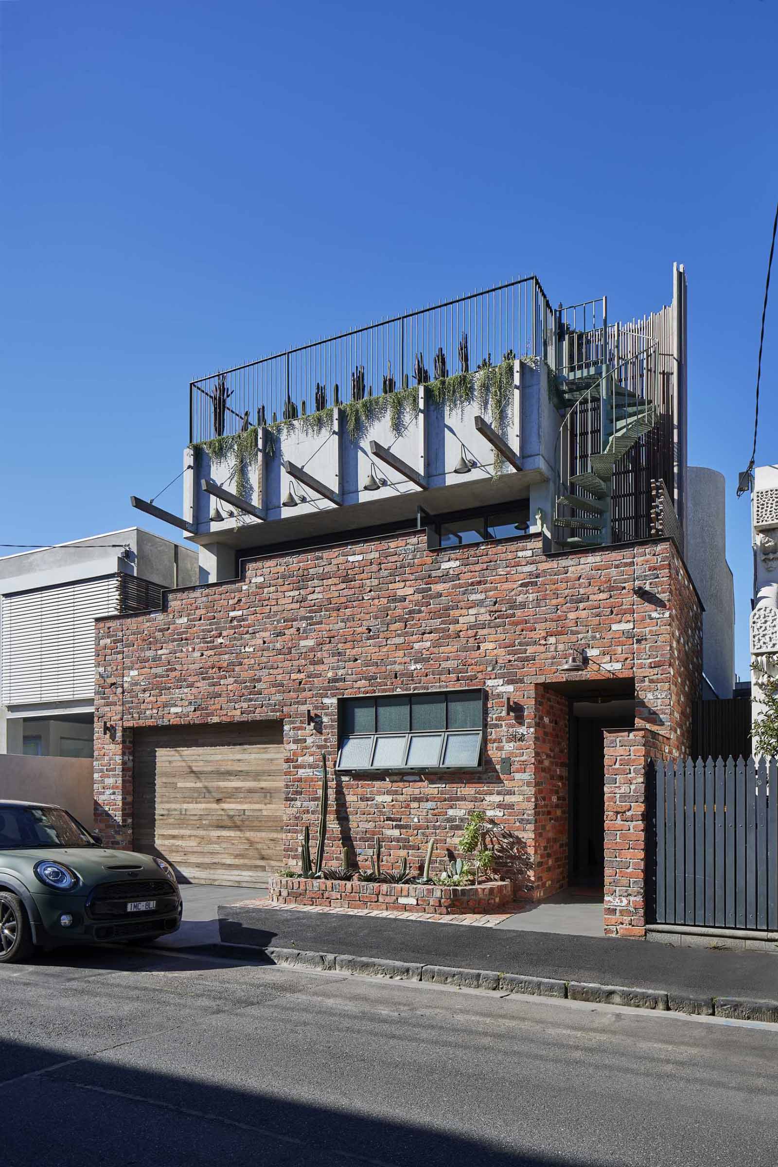 A red brick single-storey facade rises in steps towards the centre, with its pediment topped with a ribbon of steel, concealing the terrace behind. 