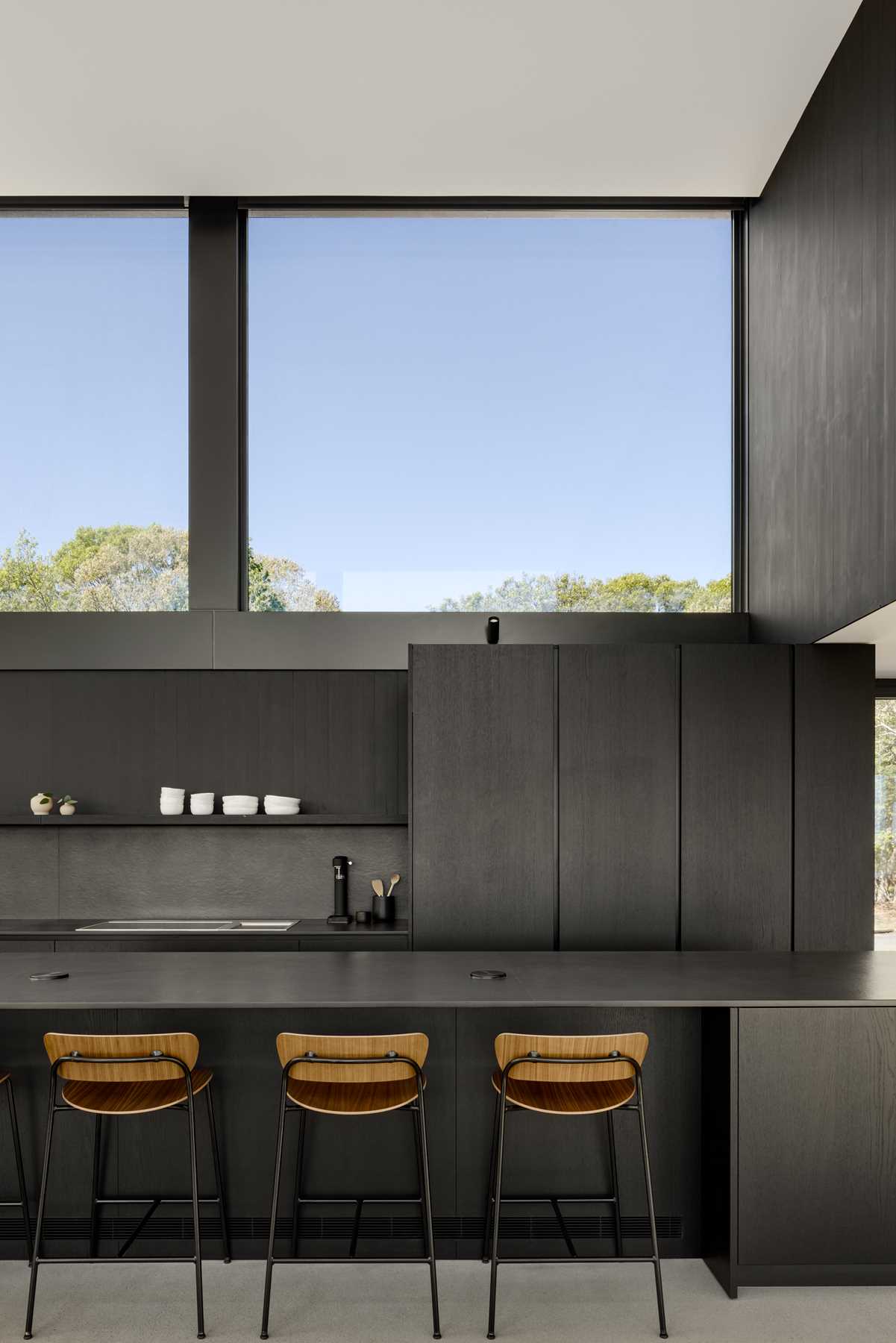 Above this modern black kitchen, large glazed openings act as a clerestory and flood the space with diffuse natural light, framing views of the surrounding treetops.