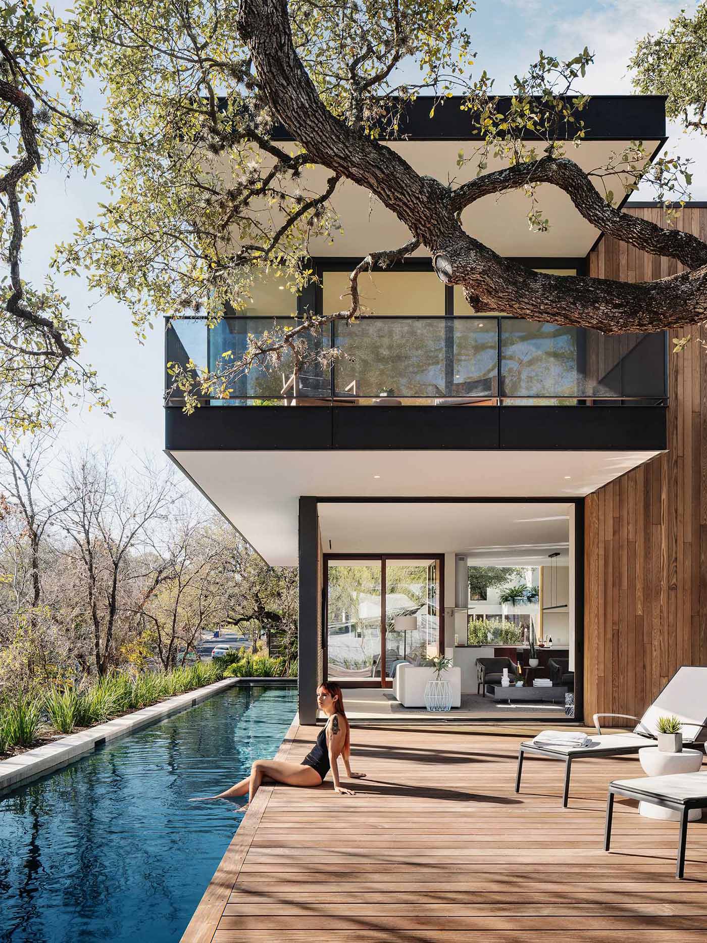 A long rectangular swimming pool runs alongside the home, while the deck is partially shaded by the large tree.