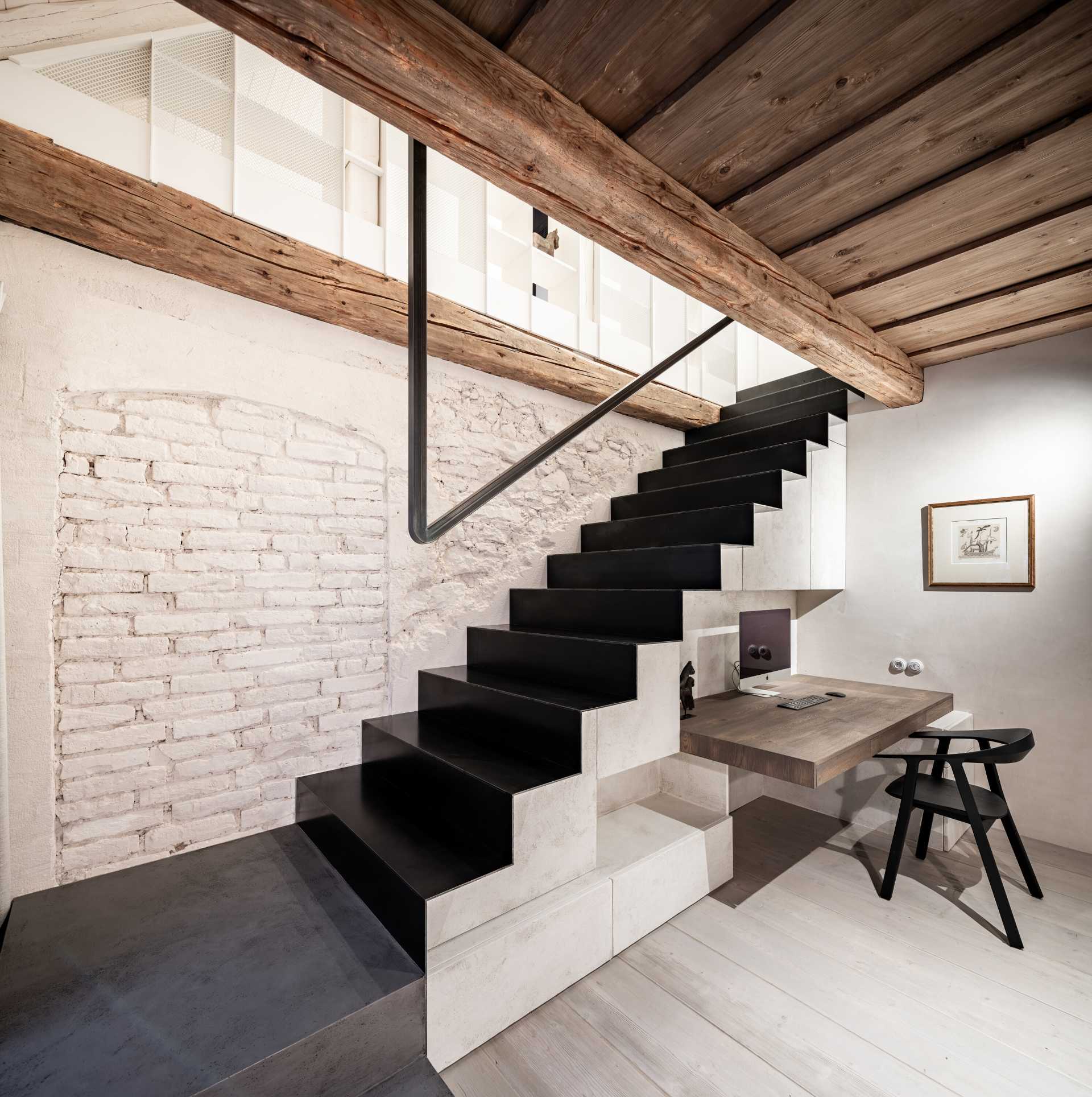 This level of the home includes a home office, with shelving included on one side of the space, while the other is dedicated to enclosed storage cabinets and a desk, and steel stairs lead up to the attic.