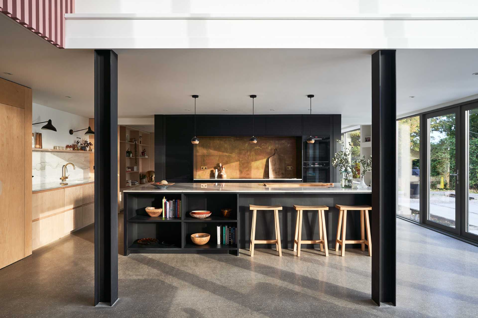 This modern black kitchen has a brass backsplash and an island with shelving for cookery book storage, while plywood has been used for the larder (pantry).