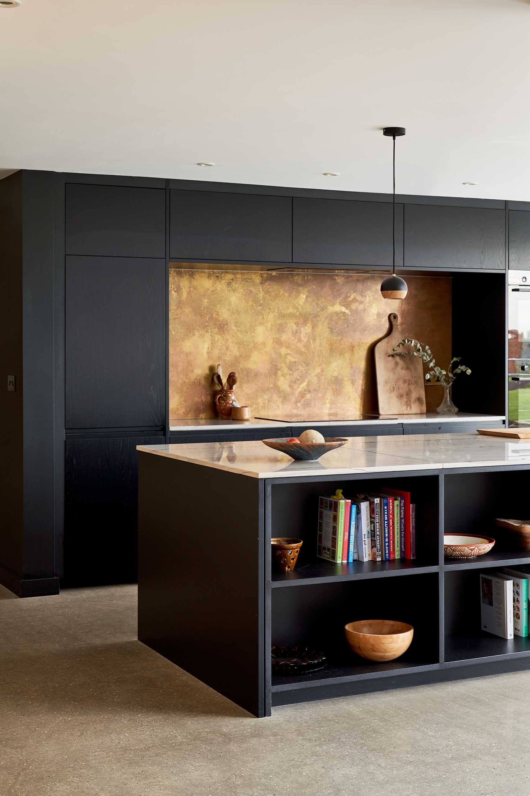 This modern black kitchen has a brass backsplash and an island with shelving for cookery book storage, while plywood has been used for the larder (pantry).