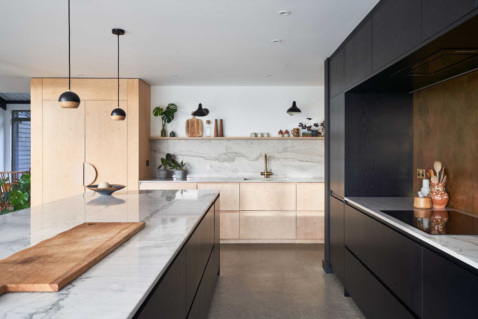 This modern black kitchen has a brass backsplash and an island with shelving for cookery book storage, while plywood has been used for the larder (pantry).